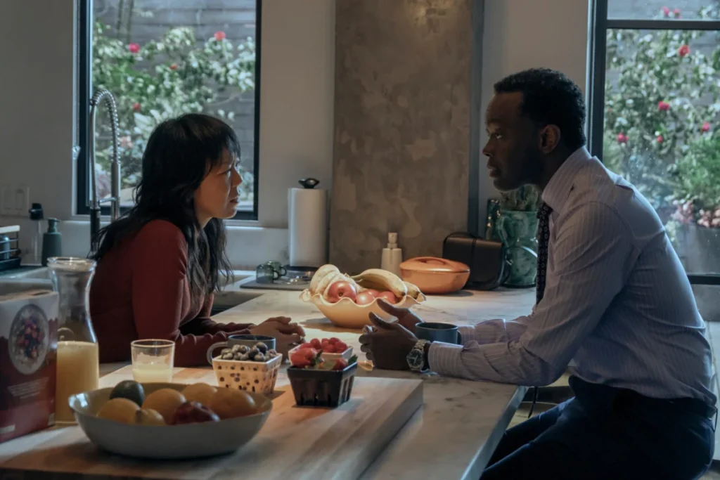 "Two people sitting at a kitchen island having a serious conversation, surrounded by fruit, juice, and soft daylight from large windows with a view of a garden outside."